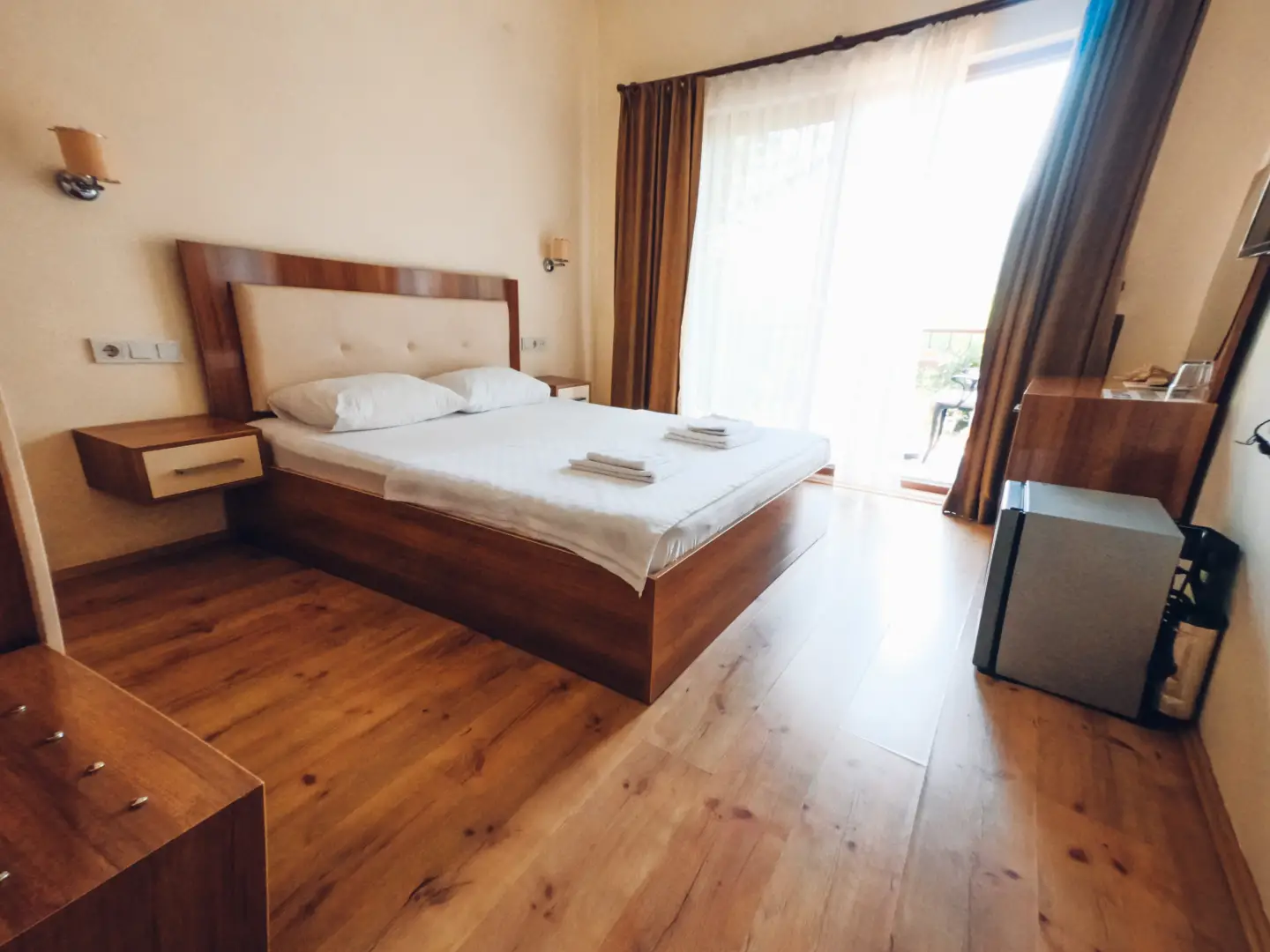 Ahşap zeminli otel odasında havlular konmuş çift kişilik yatak, mini buzdolabı ve tül perdeli balkon kapısı