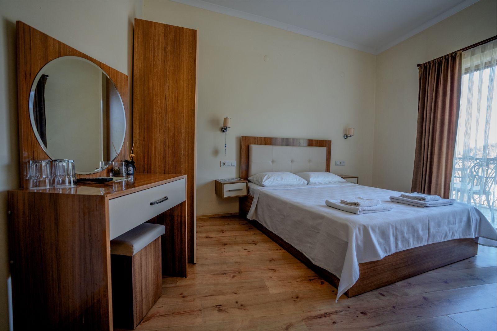 Çift kişilik yatağı, katlanmış havluları, yuvarlak aynalı ahşap makyaj masası ve balkon kapısı olan otel odası
