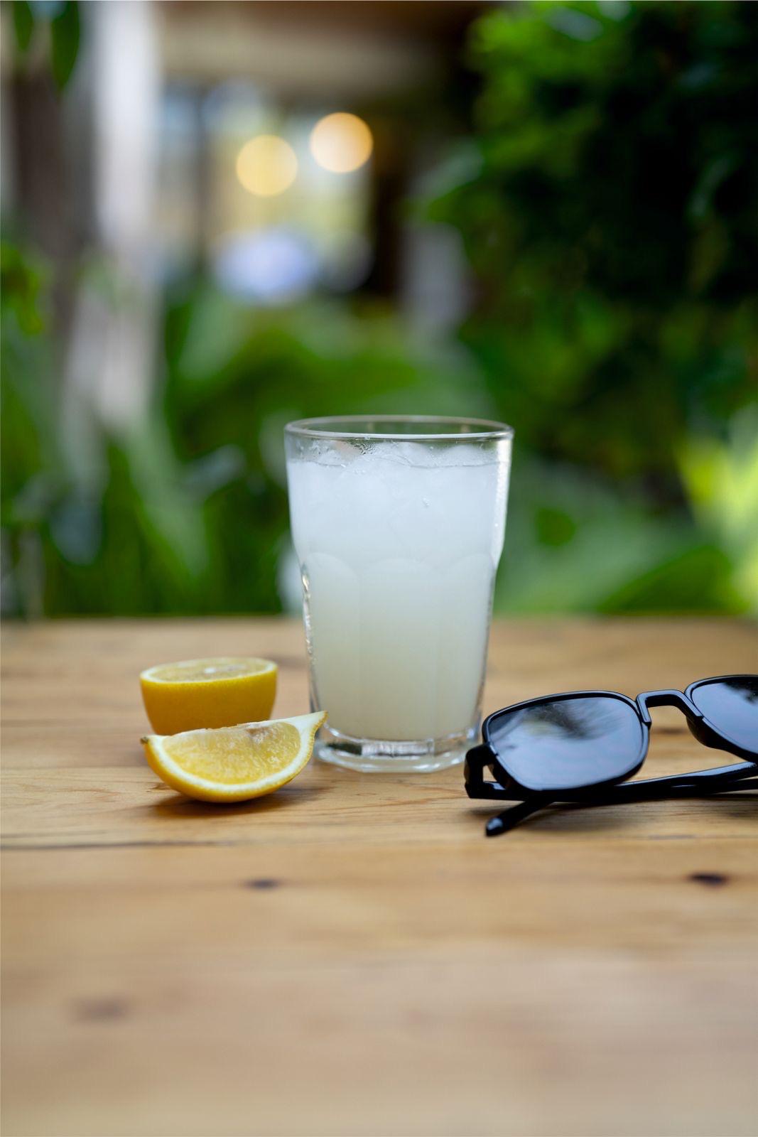 Ahşap masada buzlu limonata bardağı, yanında kesilmiş limon dilimleri ve siyah güneş gözlüğü; arkada yeşil bitkiler
