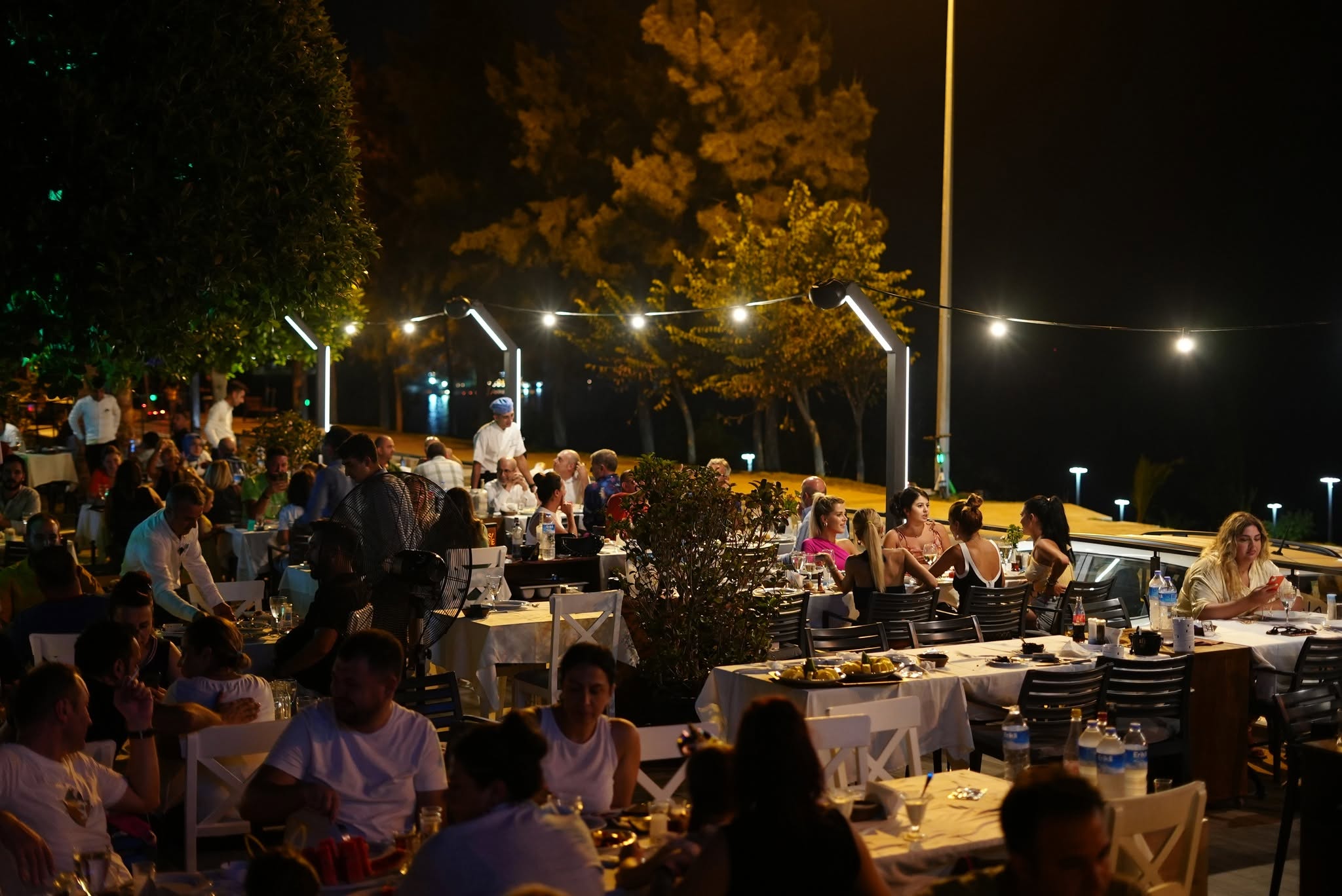 Kalamar Balık'ın açık hava bahçesinde gece, ışık zinciri altında beyaz örtülü masalarda yemek yiyen kalabalık