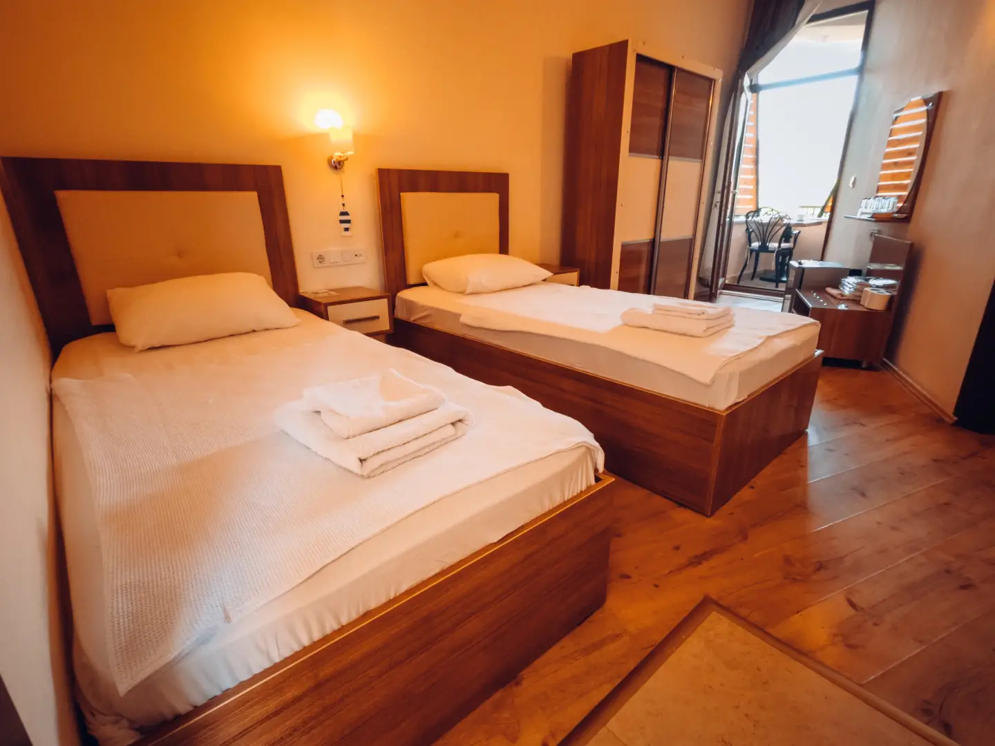 Ahşap çerçeveli iki tek kişilik yatak, katlanmış havlular, gardırop ve balkona açılan kapısı olan otel odası