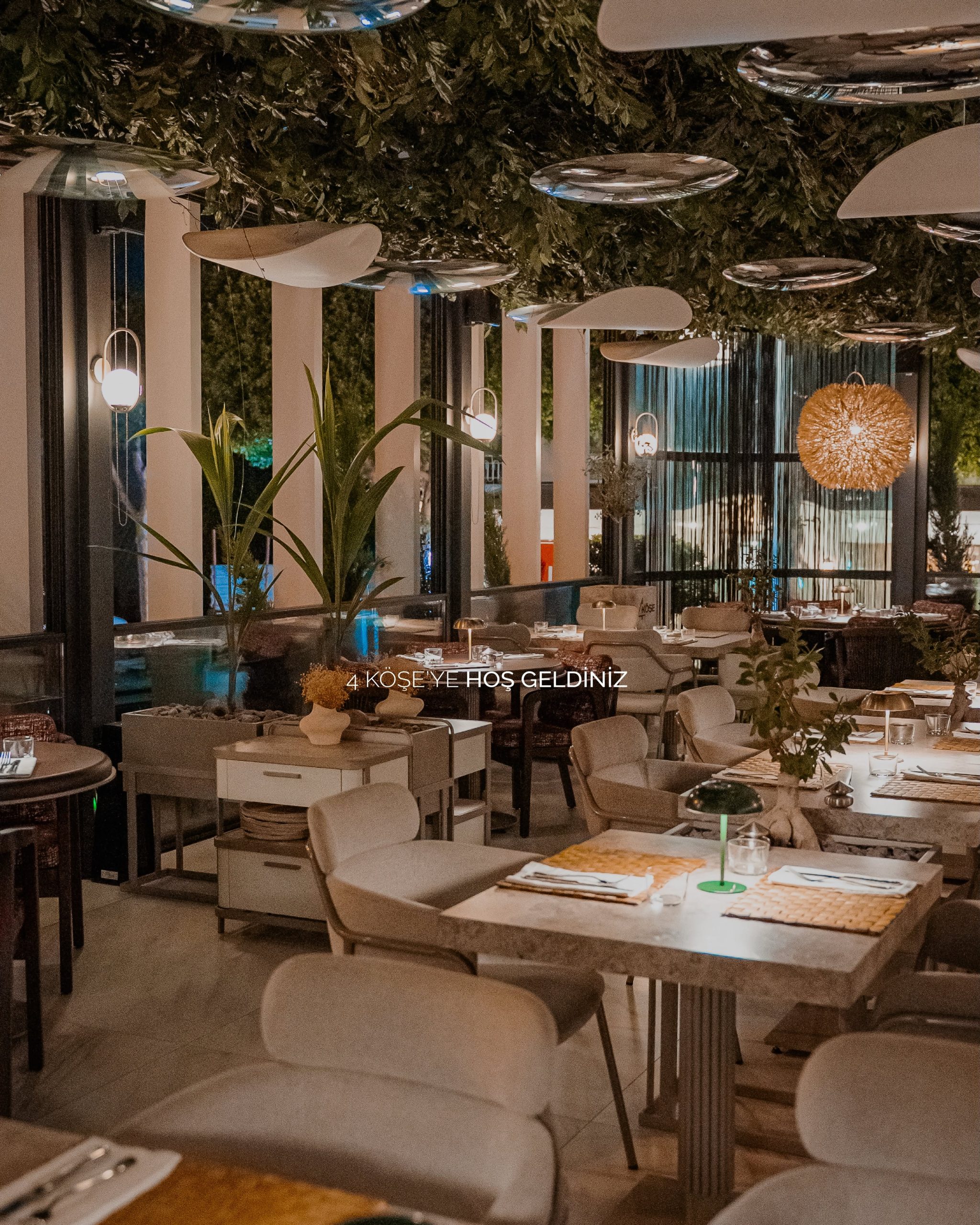 4 Köşe Restaurant'ta gece, yeşil bitkili tavan, aynalı sarkıt lambalar ve kurulu masalar; "4 Köşe'ye Hoş Geldiniz" yazısı