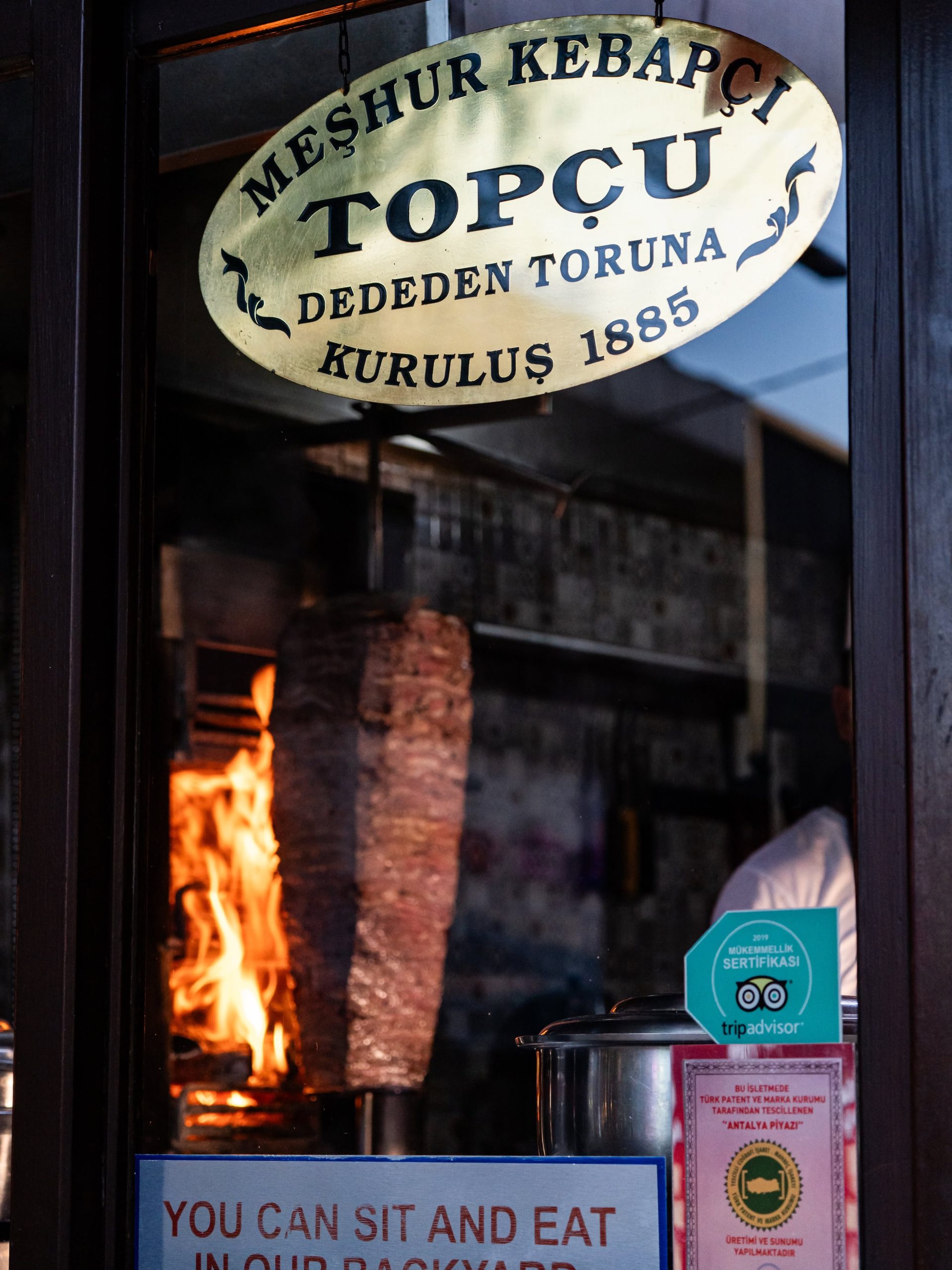 Topçu Kebap vitrininde "Meşhur Kebapçı Topçu, Kuruluş 1885" tabelası ve odun ateşi önünde dönen döner