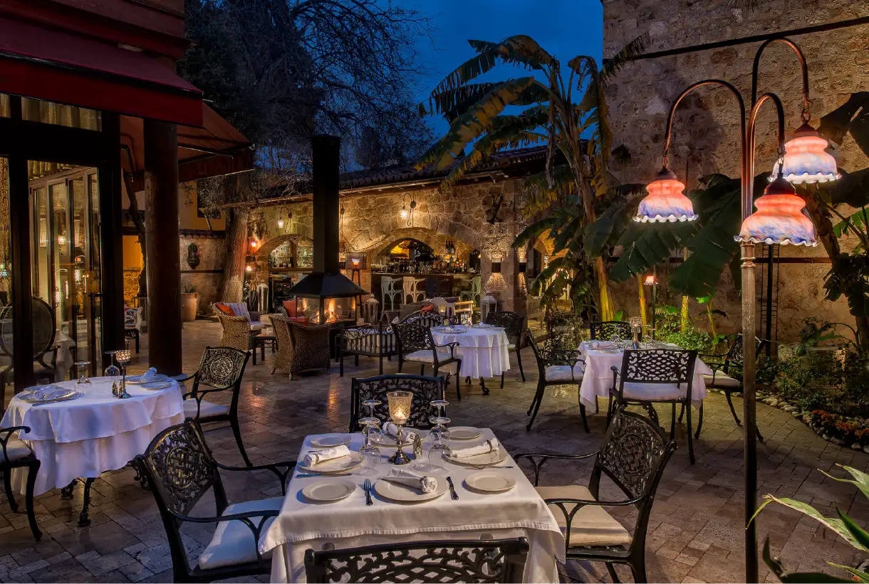 Seraser Fine Dining Restaurant'ın taş avlusunda akşam, beyaz örtülü masalar, ferforje sandalyeler, şömine ve palmiyeler