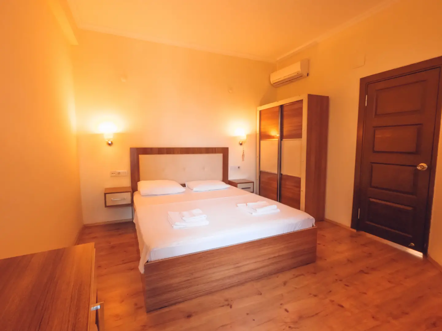 Beyaz çarşaflı çift kişilik yatak, katlı havlular, ahşap gardırop, klima ve ahşap kapılı sıcak ışıklı otel odası