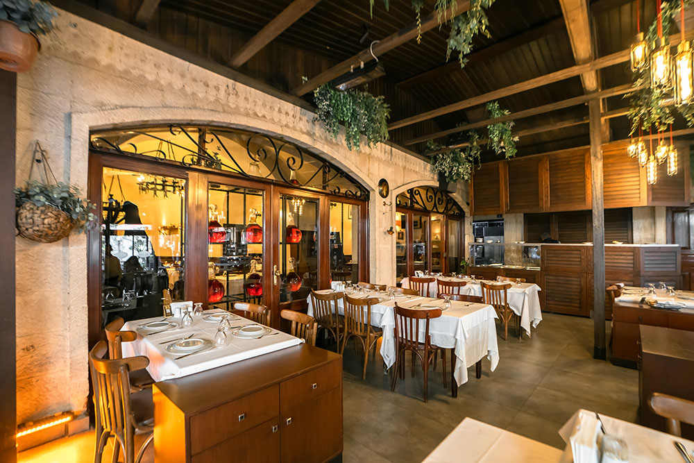 Parlak Restoran'da beyaz örtülü masalar, ahşap sandalyeler, taş kemerli camlı kapılar ve sarkan bitkilerle ahşap tavan