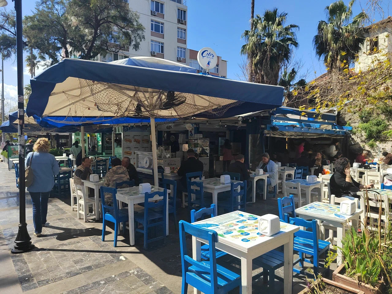 Çıtır Balık'ın açık hava bölümünde mavi sandalyeli beyaz masalar, ağlı büyük şemsiye ve palmiyeler