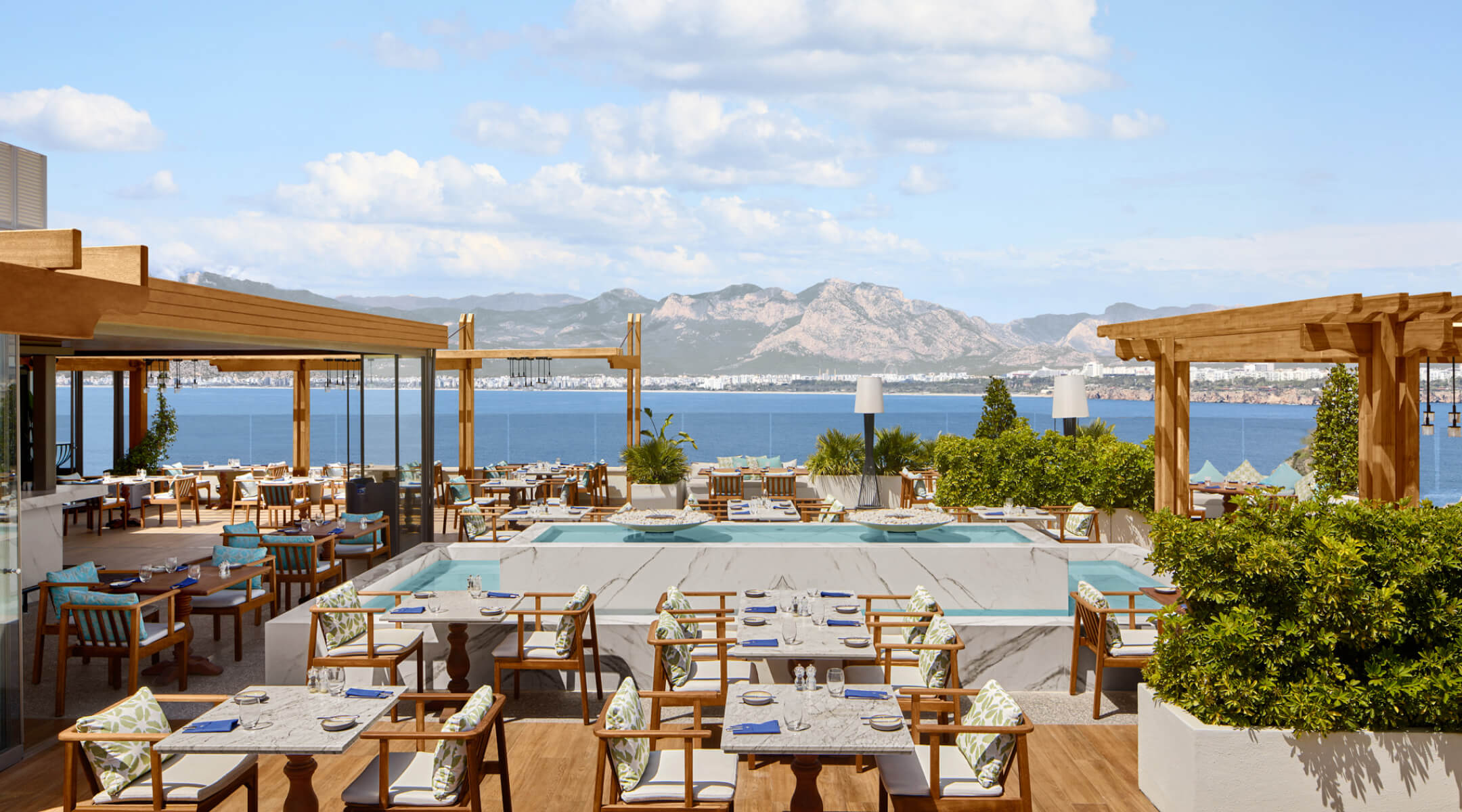 Asmani Restaurant'ın deniz manzaralı terası; ahşap masalar, havuzlu orta alan ve arkada Beydağları