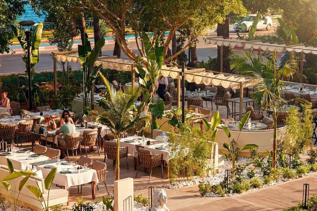Antalya Balık Evi'nin palmiyeli bahçesinde beyaz örtülü masalar, hasır sandalyeler ve tenteler; arkada yol ve deniz