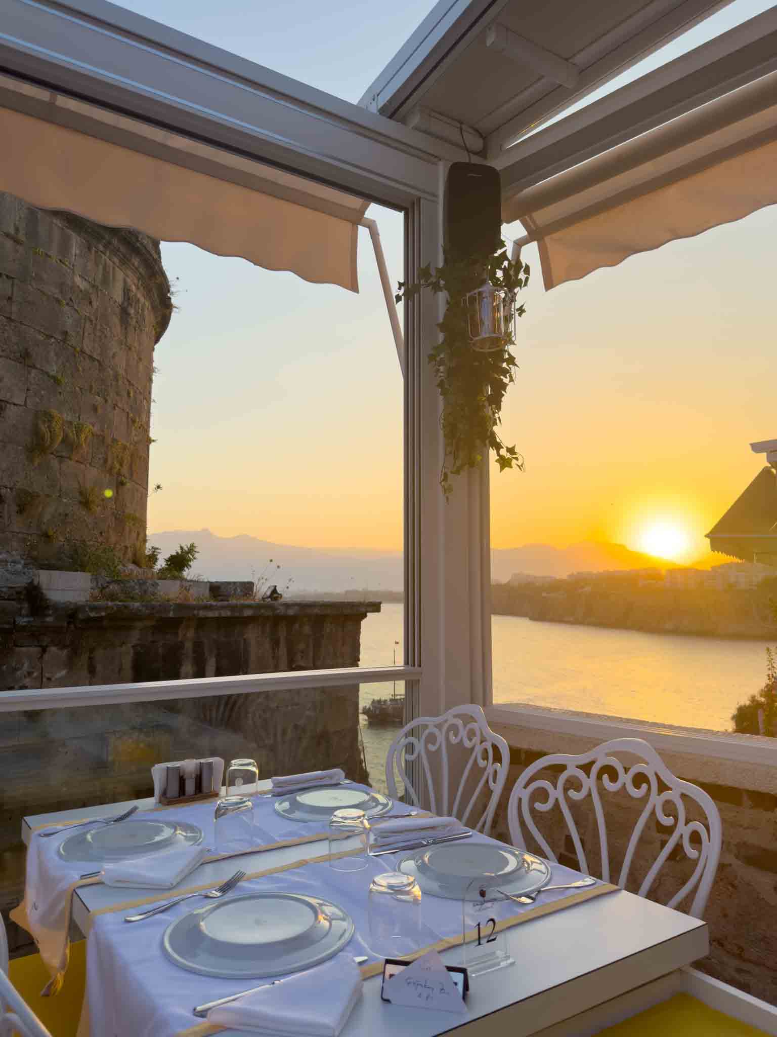 Sauvignon Restaurant terasında kurulu masa, yanında taş kule ve gün batımında deniz ile dağ manzarası