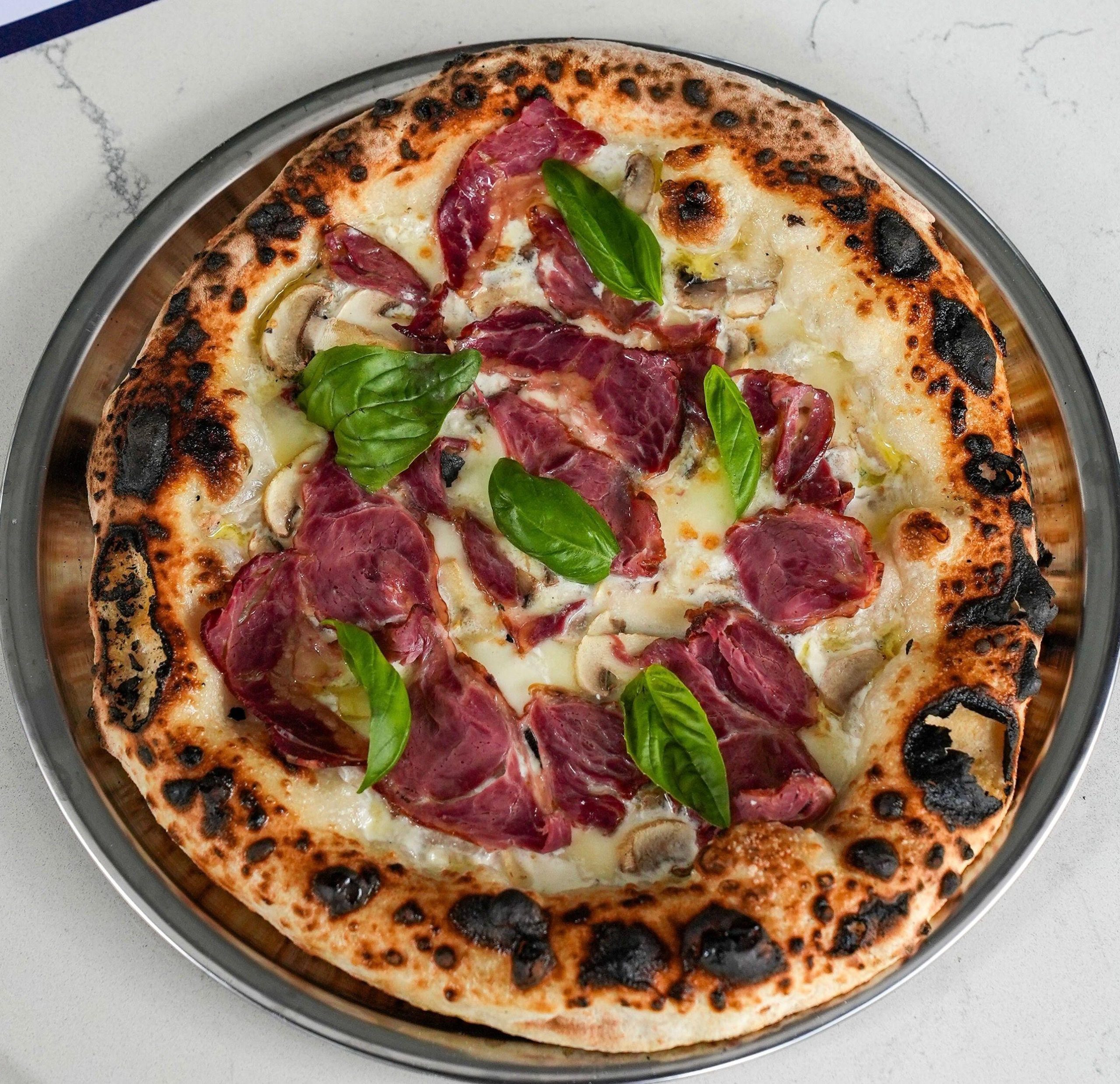 İl Vicino Pizzeria'da metal tepside kurutulmuş et, mantar, mozzarella ve fesleğenli, kenarları közlenmiş pizza