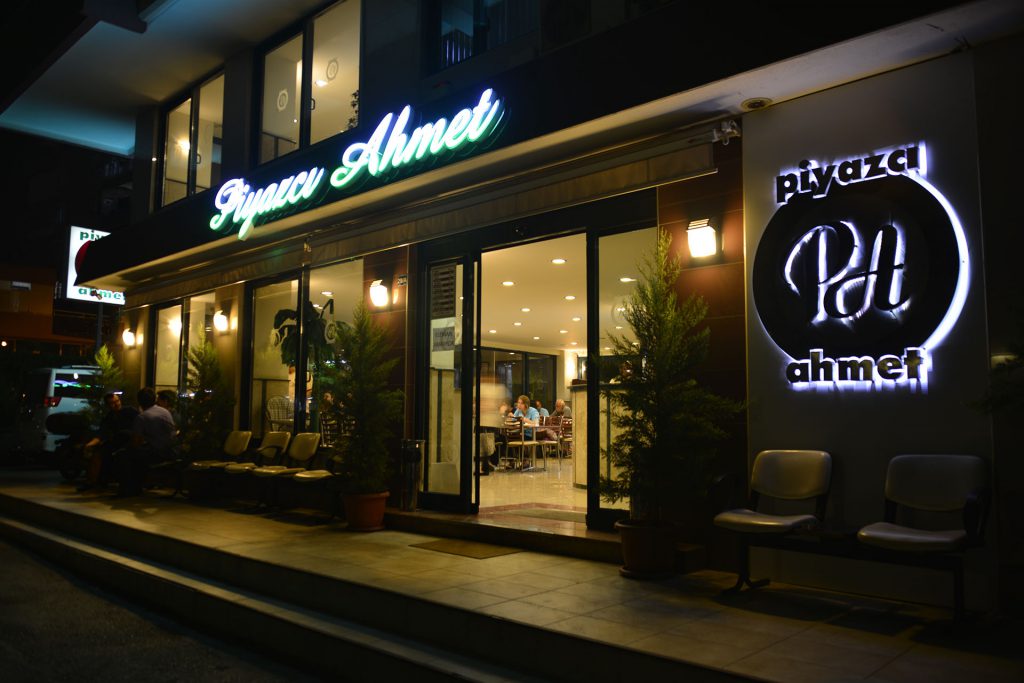 Piyazcı Ahmet'in gece ışıklı tabelası, cam girişi, içeride yemek yiyenler ve dışarıdaki bekleme sandalyeleri