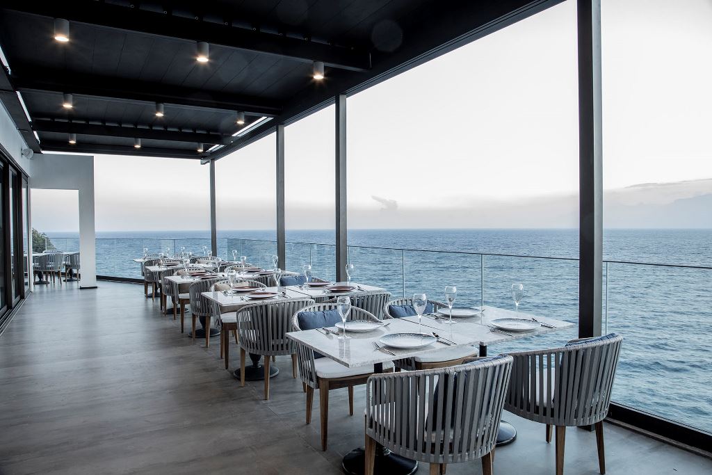 Lanthe Restaurant'ın denize bakan cam korkuluklu terası, mermer masalar ve gri örgü sandalyeler