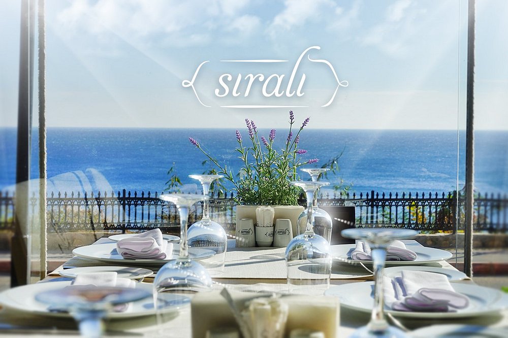 Sıralı Kebap'ta deniz manzaralı, lavanta süslü kurulu masa; ters kadehler, beyaz tabaklar ve camda Sıralı logosu