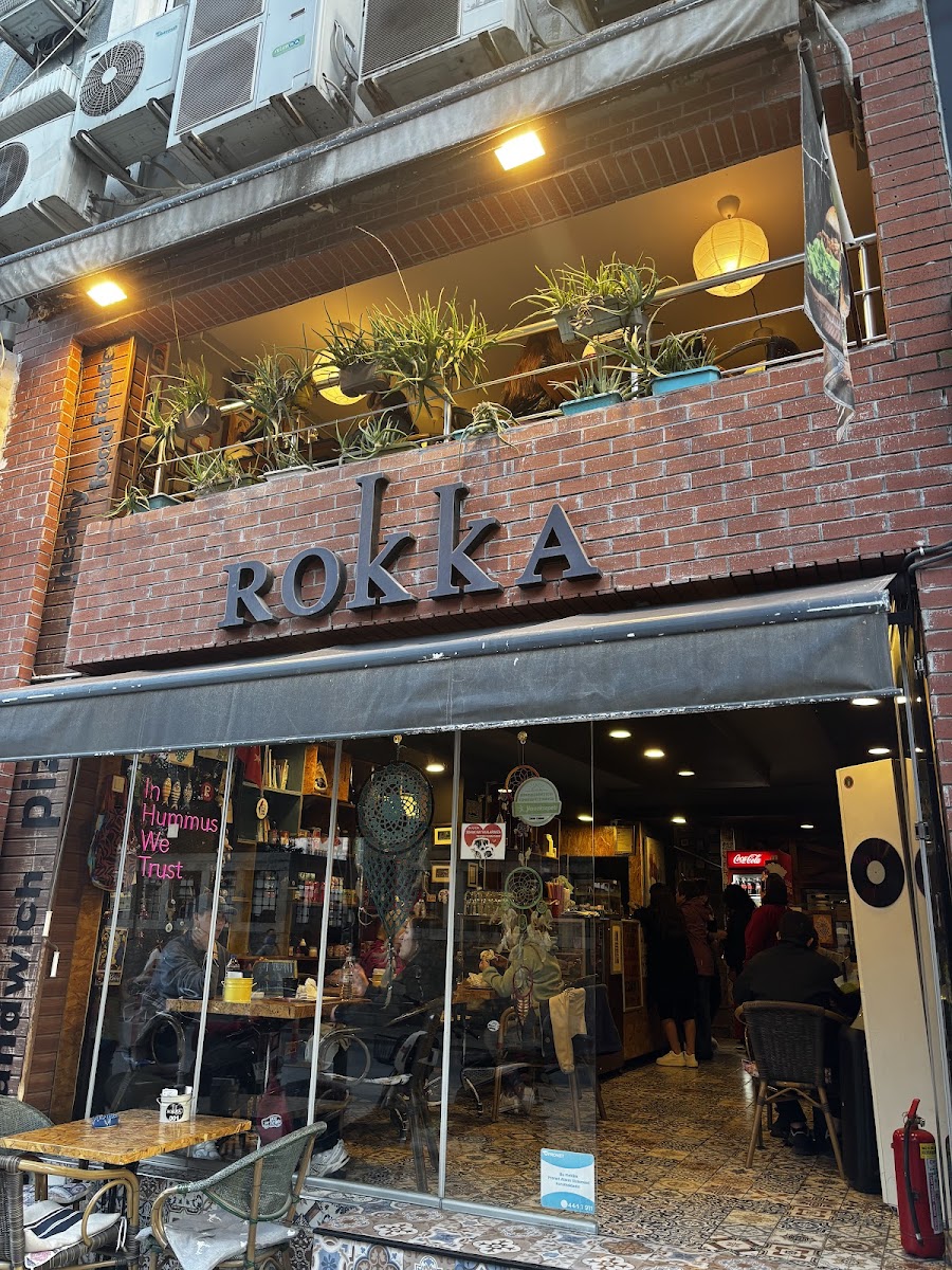 Rokka Pizza Falafel'in tuğla cephesi, ROKKA tabelası, saksı bitkili balkon ve içeride oturan müşteriler