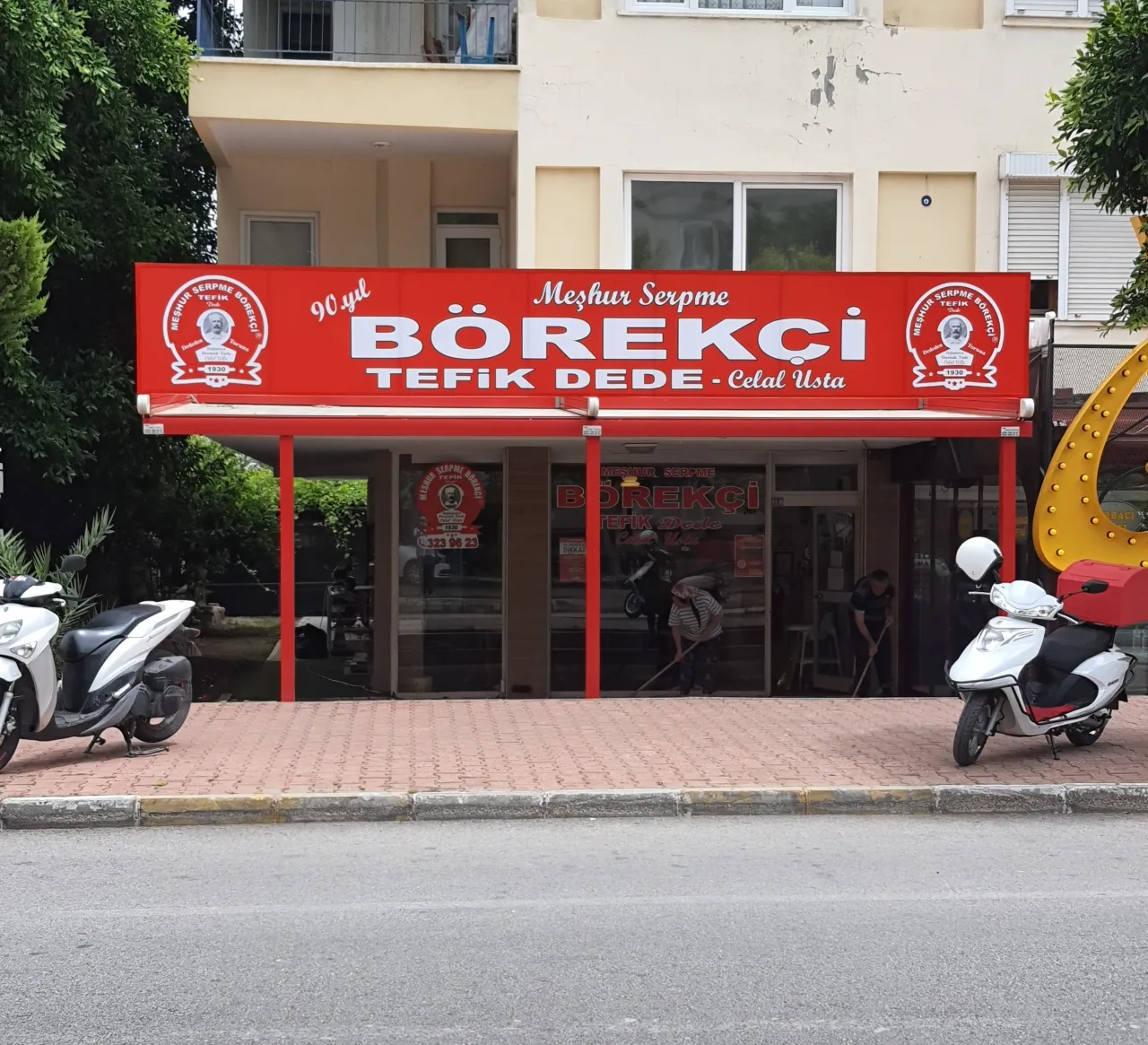 Börekçi Tevfik'in kırmızı tabelalı dükkân cephesi; tabelada "Meşhur Serpme Börekçi Tefik Dede", önünde iki scooter